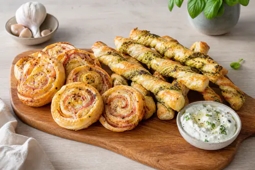 Złociste ślimaczki z ciasta francuskiego z szynką i serem oraz skręcone paluszki z pesto na drewnianej desce